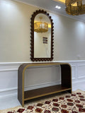 Decorative mirror with wooden frame above a console table in a room with floral rug and chandelier.